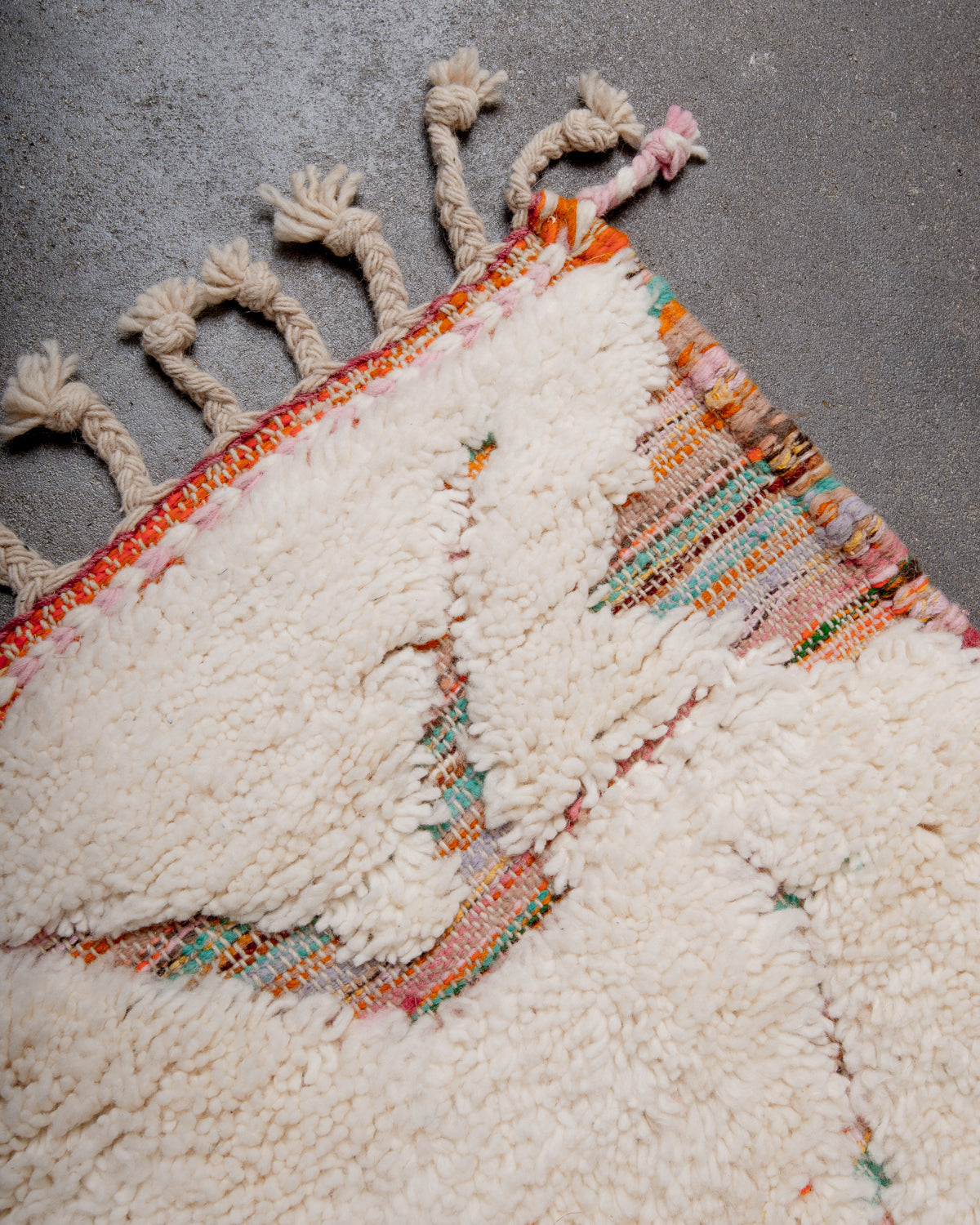 Corner of cream Beniourain wool rug with a multicoloured base and natural wool tassels laying on concrete floor.